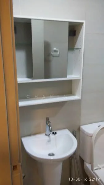 Bathroom features: white ceramic sink, chrome basin mixer faucet, wall-mounted mirror, white toilet, built-in shelving unit, light-colored tiled walls, tile flooring, glass shelf above sink; style: modern.