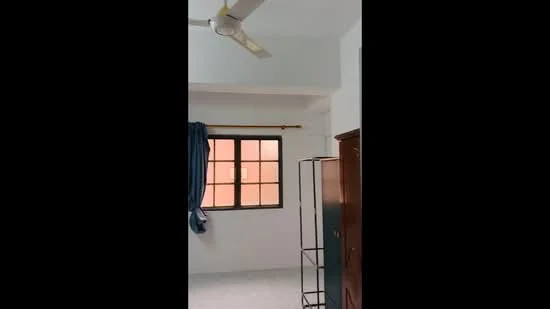 Room features: white painted walls, ceiling fan, window with grid pattern, curtains, light-colored flooring, wooden wardrobe, corner shelving unit.