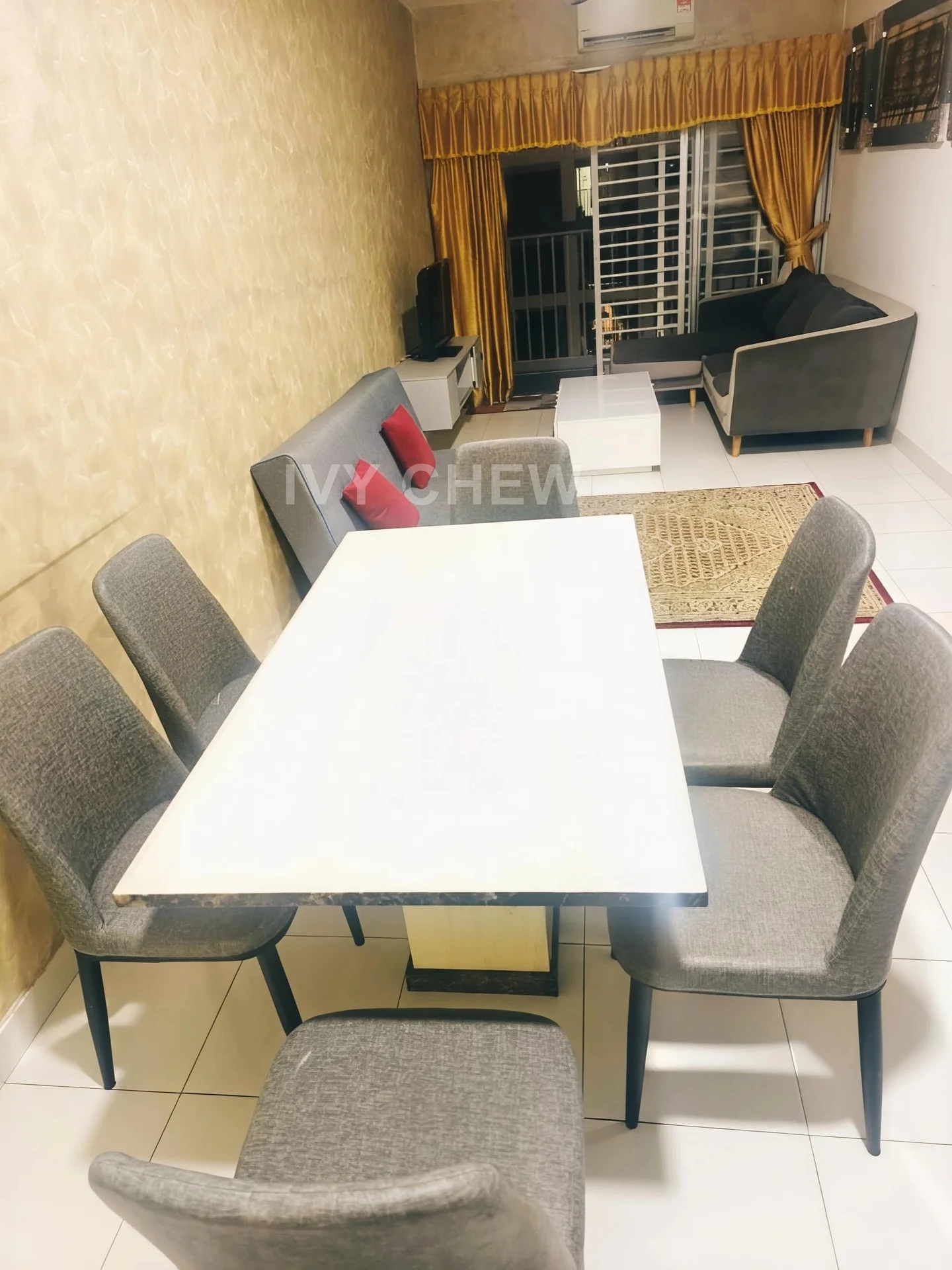 Dining features: rectangular dining table with white top, 6 upholstered dining chairs in grey, cream tiled flooring, golden wallpaper with textured pattern, wall-mounted TV, curtains in gold, sofa in grey with red accent pillows, area rug with geometric pattern, sliding glass door leading to balcony, air conditioning unit on wall; style: modern; view: city.