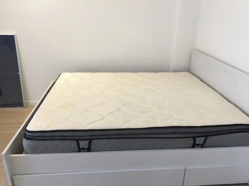 Bedroom features: queen-size bed, mattress with quilted cover, light wood bed frame, light wood flooring, white painted walls, minimalist design; style: modern.