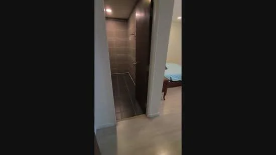 Room features: tile flooring, neutral wall colour, doorway leading to bathroom, light fixtures in ceiling, visible bed in adjacent room, wooden bed frame, blue bedding; style: modern.