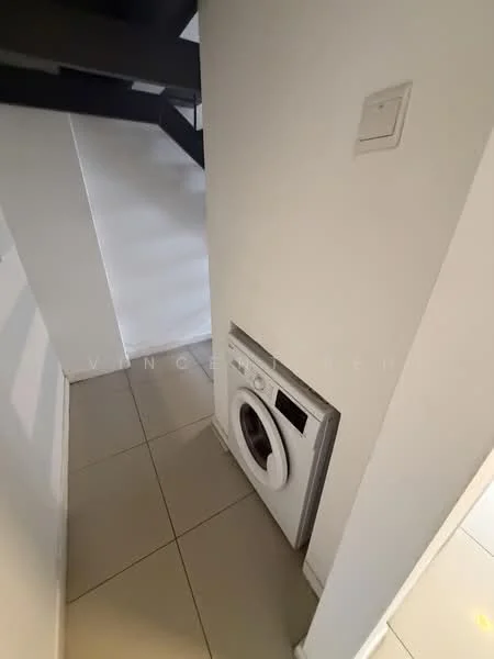 Laundry features: front-load washing machine, white walls, grey tiled flooring, light from nearby window, staircase visible in background, wall-mounted electrical switch.