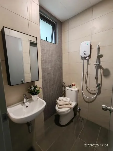 Bathroom features: grey tiled walls, light grey tiled flooring, white wall-mounted sink, chrome basin mixer faucet, one-piece toilet, shower with handheld showerhead, wall-mounted water heater, mirror with black frame, small potted plant, towels neatly arranged; style: modern.