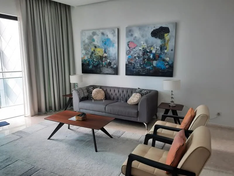 Living features: grey tufted fabric sofa, 2 beige armchairs, wooden coffee table, area rug, framed abstract artwork on walls, floor-to-ceiling curtains, sliding glass door, lamps on side tables, light-colored marble flooring; style: modern.