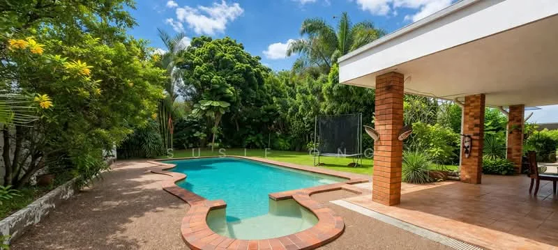 Exterior type: detached bungalow; features: swimming pool with curved edge, brick pool deck, manicured lawn, tropical landscaping with palm trees, covered patio area, exposed brick columns, surrounding greenery, outdoor dining area, sunny blue sky; style: tropical-modern.