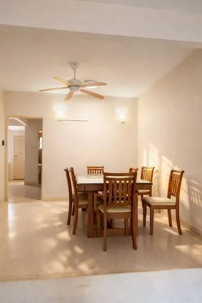 Dining features: wooden dining table, 6 wooden dining chairs with cushions, 5-blade ceiling fan, cream painted walls, marble flooring, wall-mounted light fixtures, natural light from windows, neutral decor; style: minimalist.
