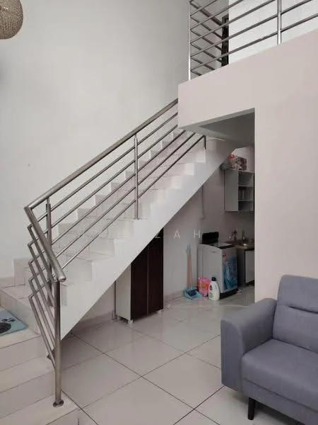 Room features: white painted walls, white tiled flooring, stainless-steel railing, staircase with white steps, grey fabric sofa, small kitchen area with cabinets, refrigerator, cleaning supplies visible, open space under staircase; style: modern.