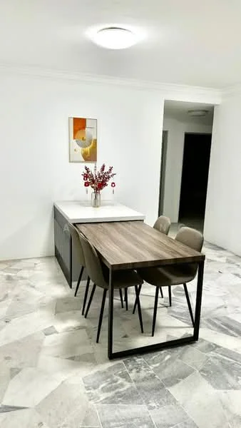 Dining features: white marble flooring, white painted walls, ceiling light, dining table with wooden top, 4 dining chairs with upholstered seats, decorative vase with flowers, framed artwork on wall, kitchen island with white countertop; style: modern.