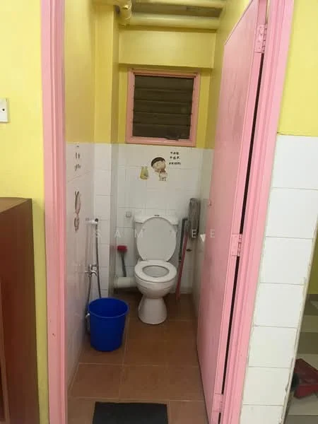 Bathroom features: white tiled walls, yellow painted walls, brown tiled flooring, one-piece toilet, sink with chrome faucet, window for ventilation, blue plastic bucket, floor mat, toilet paper holder.