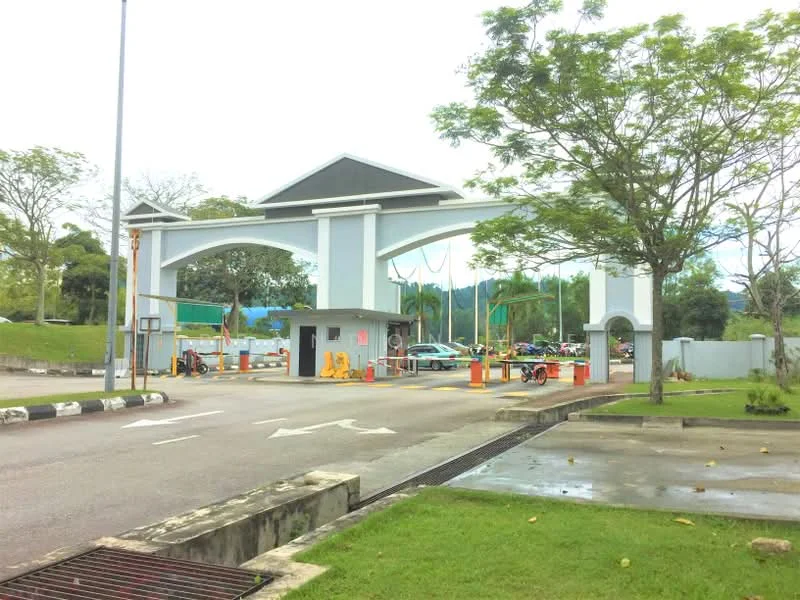 Exterior type: main entrance gate; features: arched entrance structure, grey painted facade, security booth, traffic barriers, paved driveway, landscaped greenery, surrounding trees, visible parking area; style: modern.