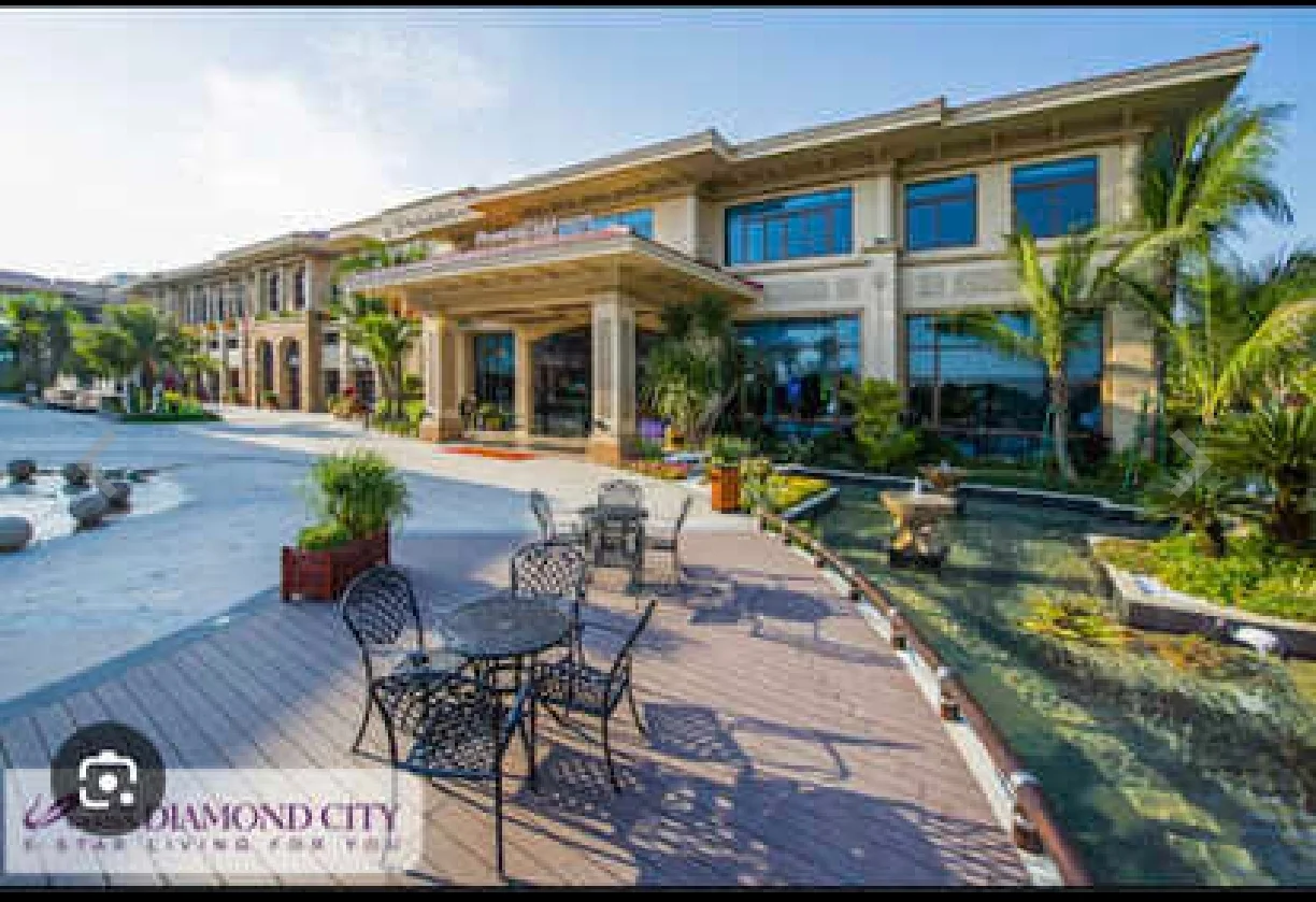 Exterior type: clubhouse; features: large entrance with columns, expansive glass windows, manicured landscaping, paved walkway, outdoor seating area with tables and chairs, water feature with pond, tropical plants, decorative stone elements; style: modern.