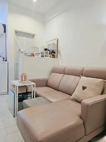 Living features: light beige leather sectional sofa, decorative throw pillow with heart design, glass side table, white walls, tile flooring, natural light from window, framed photographs on display, white cabinetry, minimalist decor; style: modern.