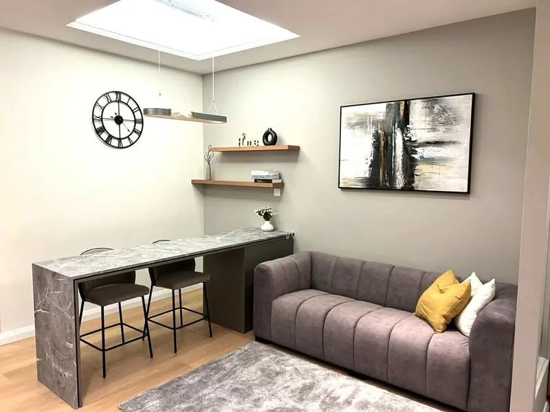 Living features: grey fabric sofa with tufted design, yellow and white decorative pillows, marble-top dining table, two black metal bar stools, wooden wall shelf, framed abstract artwork, wall clock, recessed ceiling lights, skylight, light wood flooring, neutral painted walls, area rug with soft texture; style: modern.