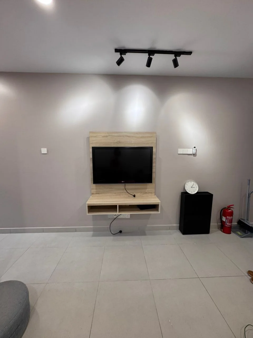 Living features: light grey painted walls, recessed lighting, wall-mounted TV, wooden TV console, grey floor tiles, black storage unit, clock on the wall, fire extinguisher, open floor space; style: modern.