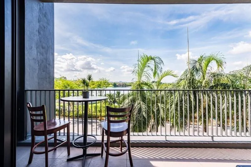Balcony features: metal railing, outdoor table, two wooden chairs, potted plant, tile flooring, view of greenery, view of water, sunny atmosphere; style: modern; view: garden.
