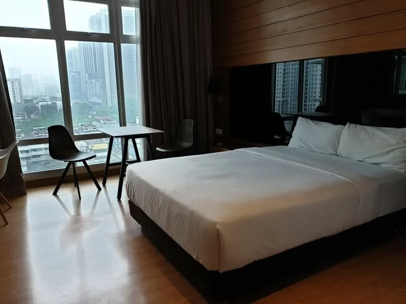 Bedroom features: king-size bed, white bedding, wooden headboard, floor-to-ceiling windows, city view, wood paneling on walls, laminate flooring, black bedside table, desk with two chairs, curtains, wall-mounted light fixture; style: modern; view: city.