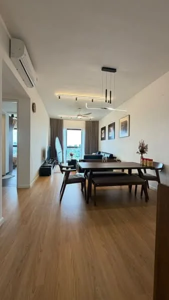 Dining features: wooden dining table, 4 dining chairs, floor-to-ceiling windows, curtains, light wood flooring, recessed LED downlights, wall-mounted TV, sofa in background, decorative vase on table, artwork on walls, air conditioning unit; style: modern; view: city. Living features: sofa, coffee table, TV console, area rug, floor-to-ceiling windows, curtains, light wood flooring; style: modern; view: city.
