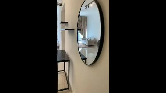 Room features: round mirror with black frame, console table with black surface, white painted walls, light-colored flooring, curtains in a neutral color, sofa visible in the background, natural light from window, shelves mounted on the wall; style: modern.