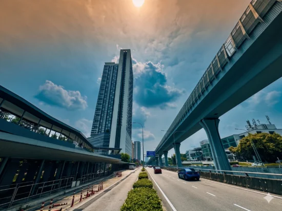 Exterior type: high-rise condominium; features: glass curtain wall, approximately 30-storey high-rise, surrounded by mature greenery, elevated monorail track, wide road with multiple lanes, urban context with commercial buildings, clear blue sky, sunlight reflecting off the building facade; style: modern.