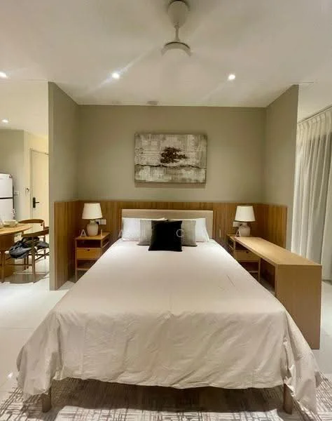 Bedroom features: king-size bed with white bedding, wooden headboard, two bedside lamps with white shades, wooden nightstands, wooden desk with chair, refrigerator visible in background, ceiling fan, wall-mounted artwork, white walls, tile flooring, curtains, natural light from window; style: modern.