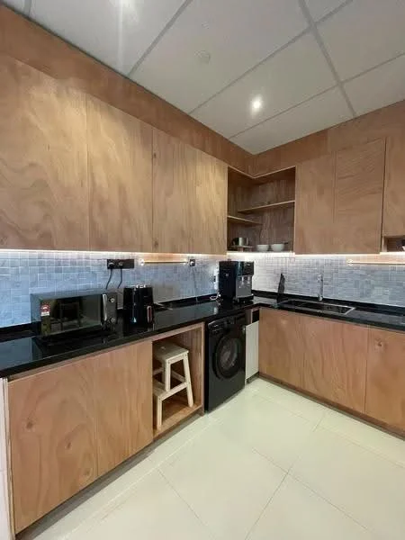 Kitchen features: wood cabinetry with natural finish, black granite countertop, white tile backsplash, built-in oven, stainless-steel fridge, black washing machine, microwave, stool, recessed lighting, under-cabinet lighting, open shelving, ceramic tile flooring; style: modern.