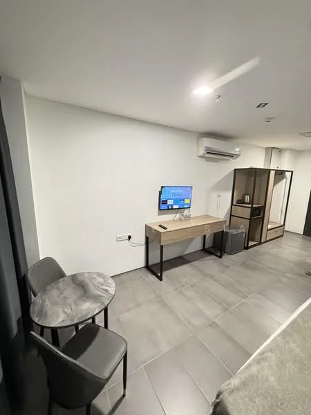 Room features: grey tiled flooring, white painted walls, wall-mounted TV, wooden desk with black metal legs, small round dining table, two grey upholstered dining chairs, mini fridge, air conditioning unit, open shelving, kitchenette area with sink; style: modern.