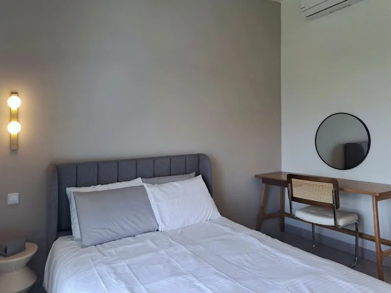 Bedroom features: king-size bed with grey upholstered headboard, white bedding, light grey painted walls, wooden desk with chair, round mirror above desk, wall-mounted sconce lighting, air conditioning unit, grey flooring; style: modern.