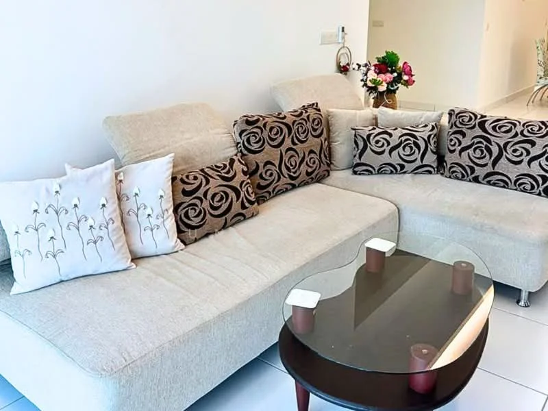 Living features: L-shape sectional sofa in beige, decorative cushions with floral pattern, glass coffee table with wooden legs, white painted walls, tile flooring, floor-to-ceiling windows, decorative flower arrangement, area rug with geometric pattern; style: modern.