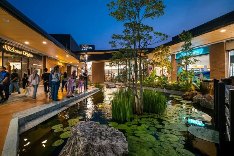 Exterior type: shopping mall; features: water feature with pond, lily pads in pond, landscaped greenery, pathway with stone edging, outdoor seating area, storefronts visible, night lighting, brick facade, modern architecture; style: contemporary.