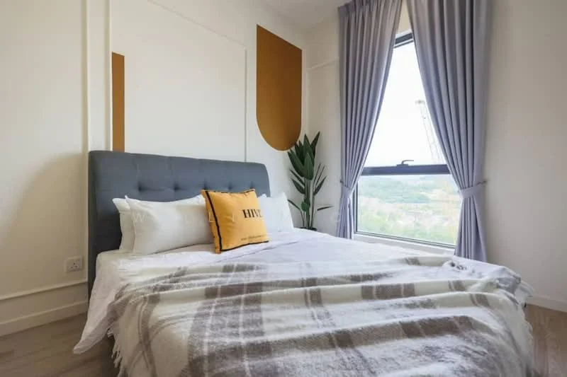 Bedroom features: king-size bed with grey upholstered headboard, white bedding with decorative pillows, plaid throw blanket, floor-to-ceiling window, curtains in light grey, indoor plant, wooden flooring, accent wall in warm tone; style: modern; view: garden.
