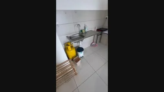 Kitchen features: stainless-steel sink, chrome faucet, white tiled walls, grey tiled flooring, gas cylinder, cleaning supplies, plastic trash bin, wooden shelving unit.