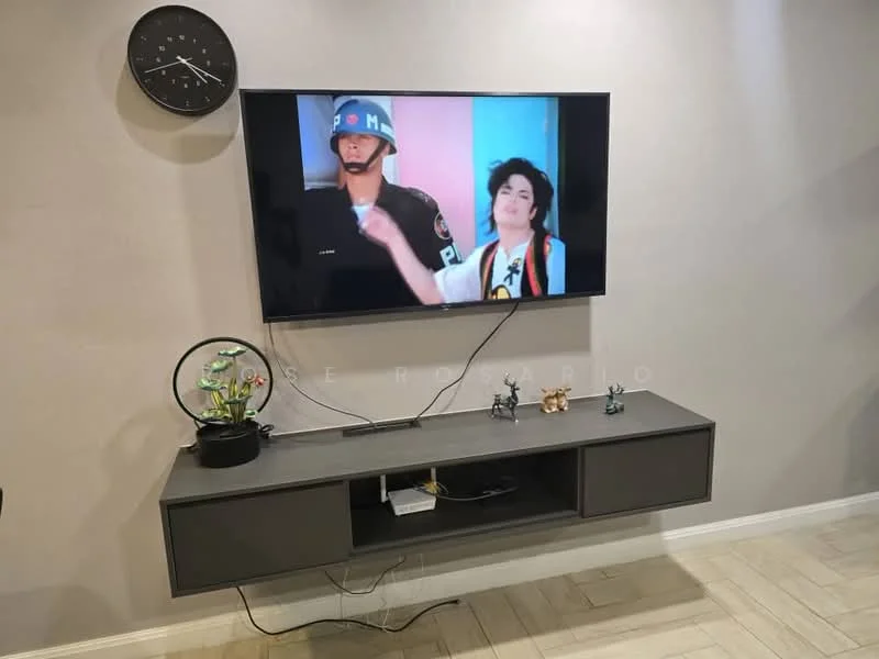 Living features: wall-mounted TV, floating TV console in grey, decorative figurines on console, wall clock, light-colored wall, wooden flooring, decorative plant in ceramic pot; style: modern.
