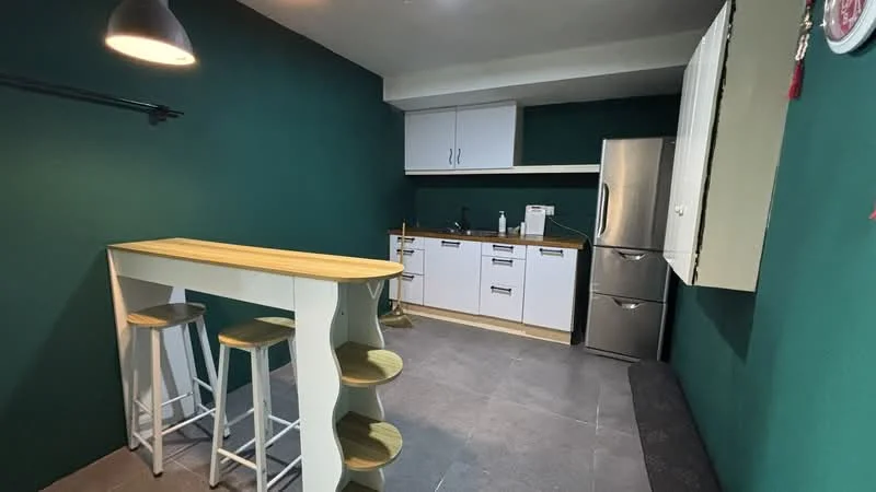 Kitchen features: green painted walls, grey tile flooring, white cabinetry, stainless-steel fridge, built-in oven, countertop with wooden finish, bar counter with stools, sink, kitchen utensils visible, light fixture, open shelving, wall-mounted storage; style: modern.