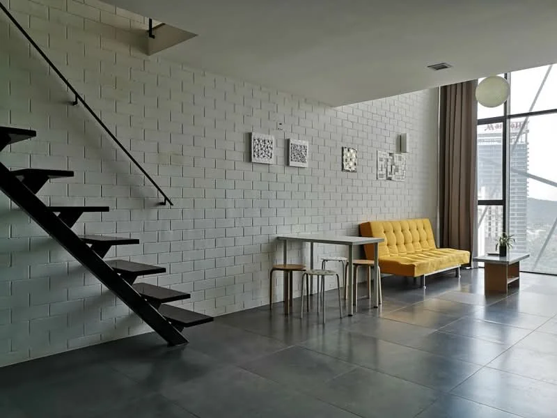 Living features: yellow fabric sofa, black metal staircase, white painted walls, grey tile flooring, glass window with city view, small wooden coffee table, two wooden stools, framed artwork on walls, hanging light fixture; style: modern; view: city.
