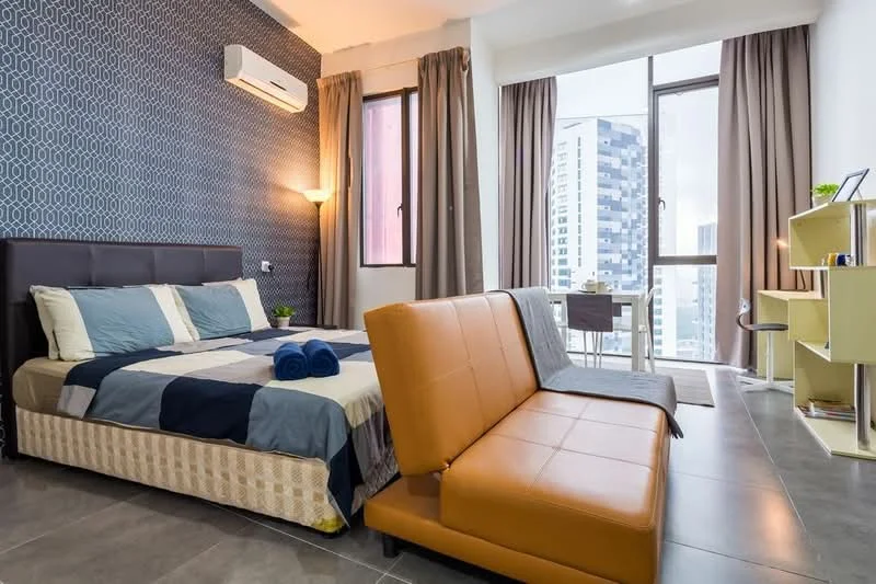 Bedroom features: king-size bed with patterned bedding, upholstered headboard, 2 blue towels on bed, floor-to-ceiling windows, grey curtains, air conditioning unit, wall-mounted light fixture, grey sofa bed, small dining table with chairs, open shelving unit, decorative plant on shelf, tile flooring; style: modern; view: city.