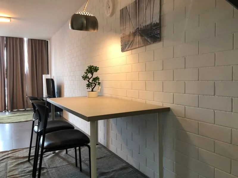 Dining features: white painted walls, concrete flooring, table with light wood top, black dining chairs, pendant light fixture, framed artwork on wall, decorative plant in pot, area rug under table; style: modern.