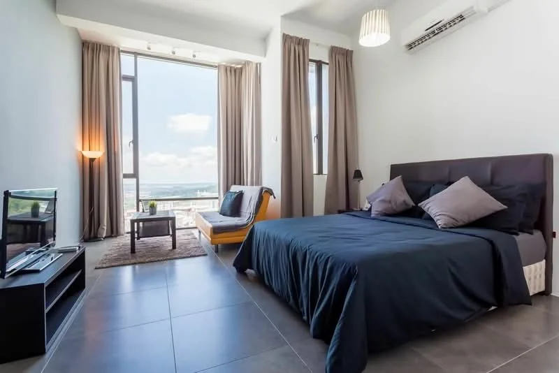 Bedroom features: king-size bed with dark bedding, upholstered headboard, floor-to-ceiling windows, sheer curtains, dark wood bedside table, table lamp, TV console with TV, area rug, sofa chair, grey tiled flooring, wall-mounted air conditioning unit, decorative wall art; style: modern; view: city.
