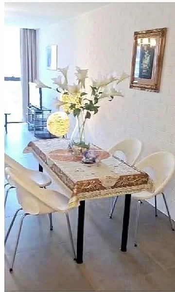 Dining features: dining table with decorative tablecloth, four white plastic dining chairs, vase with flowers, decorative round lamp, framed artwork on wall, light-colored walls, large window with sheer curtains, tile flooring; style: modern; view: city.