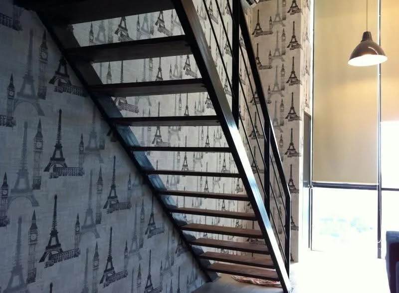 Room features: staircase with wooden steps, metal railing, wallpaper with Eiffel Tower pattern, large window providing natural light, light fixture hanging from ceiling, neutral wall color, open space design; style: modern; view: city.