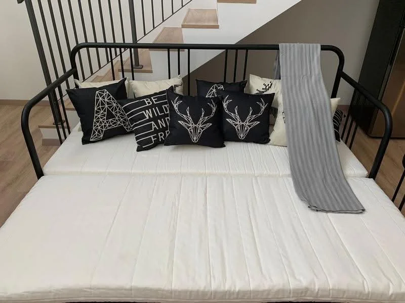 Room features: daybed with black metal frame, white mattress, decorative black and white pillows with geometric and animal designs, grey throw blanket, wooden staircase in background, metal railing, light wood flooring, neutral wall color; style: modern.