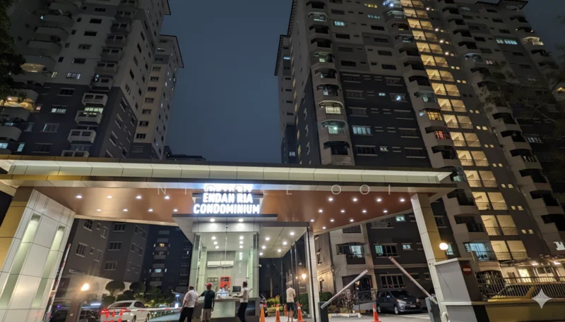 Exterior type: high-rise condominium; features: glass curtain wall, illuminated entrance signage, double-volume main lobby, covered entrance with awning, surrounded by landscaped gardens, multiple balconies on upper floors, security guard at entrance, driveway with interlocking pavers; style: modern.