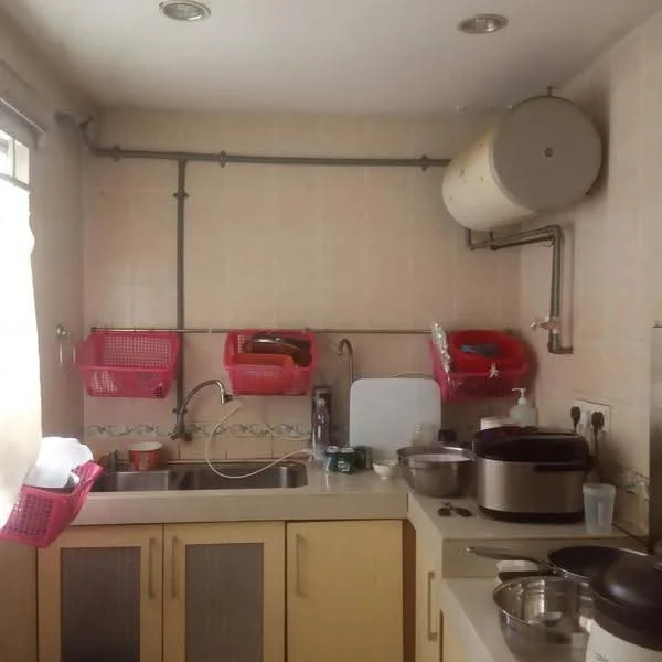 Kitchen features: single-bowl stainless-steel sink, chrome pull-down faucet, light-colored cabinetry, countertop with integrated sink, wall-mounted water heater, pink plastic storage baskets, ceramic tile backsplash, stainless-steel cooking pot, various kitchen utensils, window with sheer curtain, wall-mounted pipes, tile flooring.