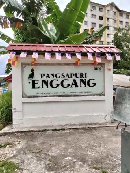 Exterior type: low-rise apartment block; features: signage with name 'Pangsapuri Enggang', decorative flags, concrete wall, roof overhang, surrounding greenery, paved ground, visible residential building in background; style: modern.