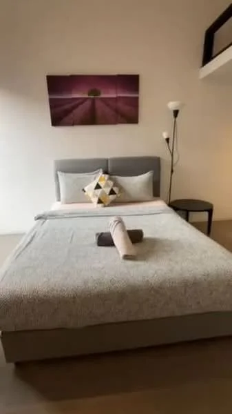 Bedroom features: queen-size bed, upholstered headboard, decorative throw pillow, bedside table, floor lamp, framed artwork on wall, light-colored bedding, textured bedspread, wooden flooring; style: modern.