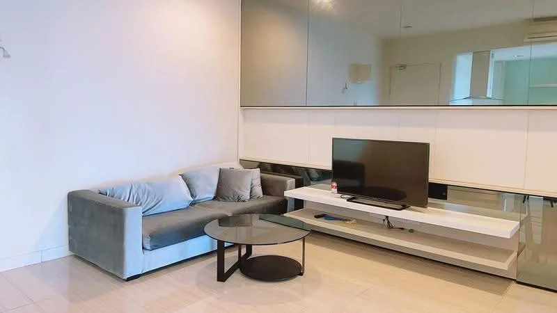 Living features: grey fabric 3-seater sofa, black round coffee table, wall-mounted TV, white TV console, glossy white wall, mirrored wall, recessed lighting, cream tiled flooring, air conditioning unit; style: modern.