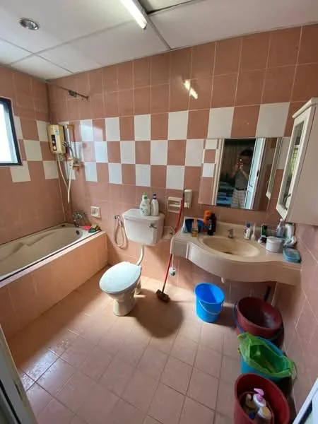 Bathroom features: pink tiled walls, white bathtub, toilet, single-sink vanity with cream countertop, mirror above vanity, showerhead, wall-mounted faucet, floor tiles, window, various toiletries on countertop, blue bucket, red bucket, green bag.