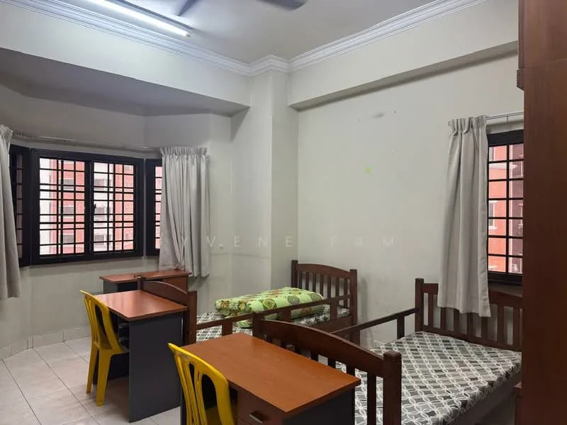 Room features: two single beds with wooden frames, two wooden study tables, yellow plastic chairs, white walls, tile flooring, two windows with black metal grills, curtains on windows, ceiling fan.