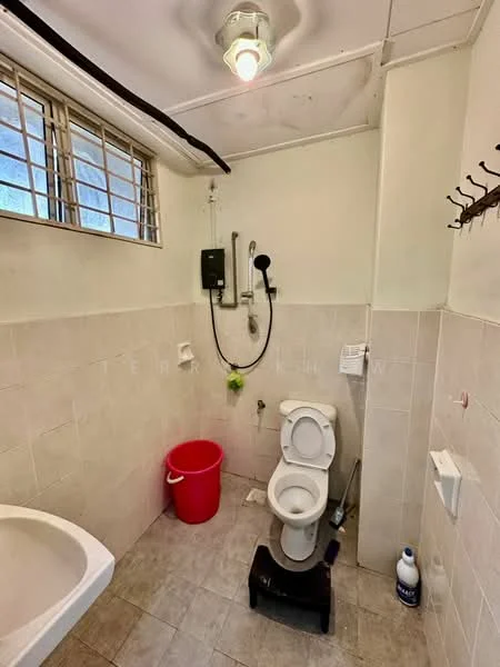 Bathroom features: white toilet, shower with handheld showerhead, wall-mounted shower control, small red bucket, white sink, tile flooring, light fixture on ceiling, window with metal grille, toilet paper holder, cleaning bottle.