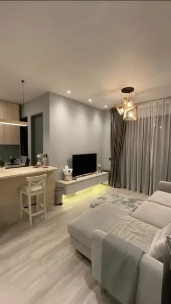 Living features: light grey fabric sectional sofa, coffee table, wall-mounted TV, TV console with LED lighting, pendant light fixture with geometric design, recessed downlights, floor-to-ceiling windows with sheer curtains, laminate flooring, open kitchen layout, bar stools at kitchen counter, neutral wall paint, decorative wall art; style: modern.