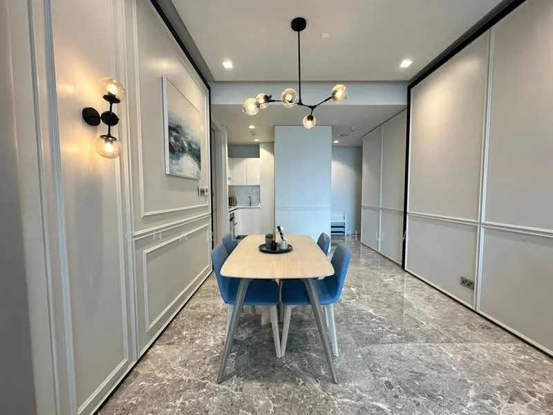 Dining features: marble flooring, white painted walls with crown moulding, 6-seater dining table, upholstered dining chairs in blue, modern chandelier with multiple bulbs, wall-mounted sconce light, open kitchen view, recessed downlights; style: modern. Kitchen features: white cabinetry, stainless-steel appliances, open layout to dining area; style: modern.